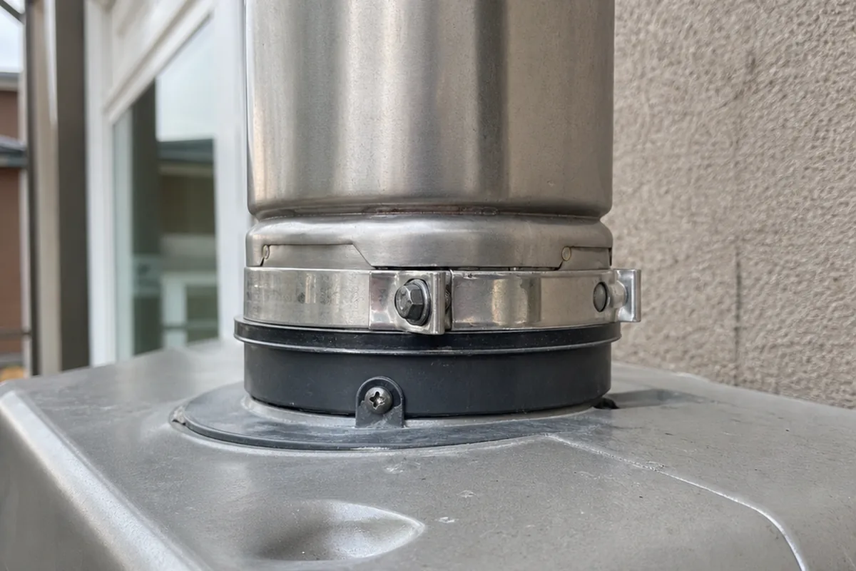 Close macro of the stainless steel vent connection at the top of a compact tankless water heater, showing the clamp band and seal against a nearby wall