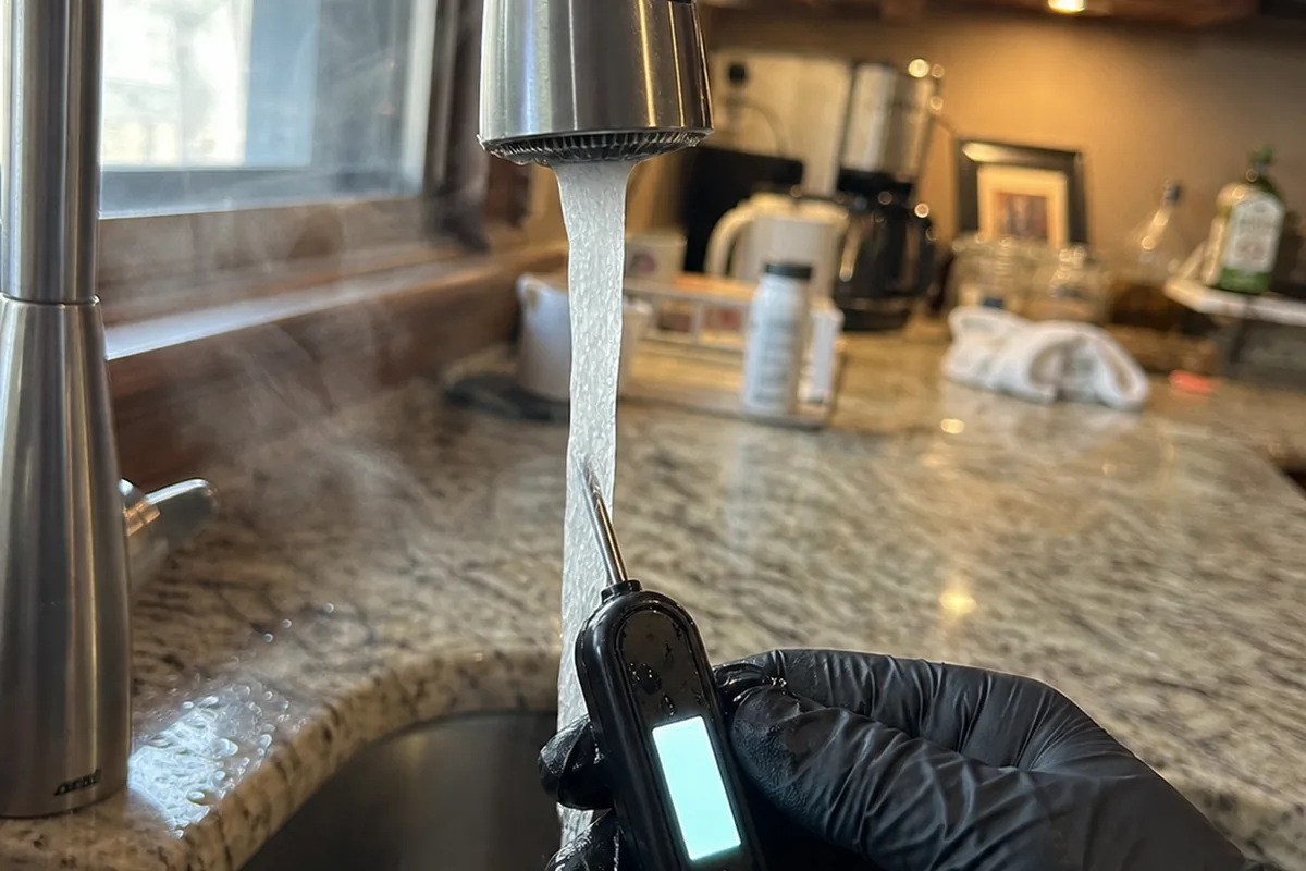 A gloved hand holding a temperature probe under a running kitchen faucet in a Rancho Santa Fe estate, checking hot water delivery temperature during an inspection