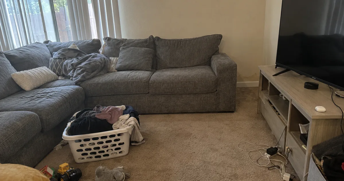 A lived-in rental living room with a gray sectional sofa pushed against the wall, a laundry basket and toys on the carpet, and a faint water stain at the baseboard just past the sofa arm