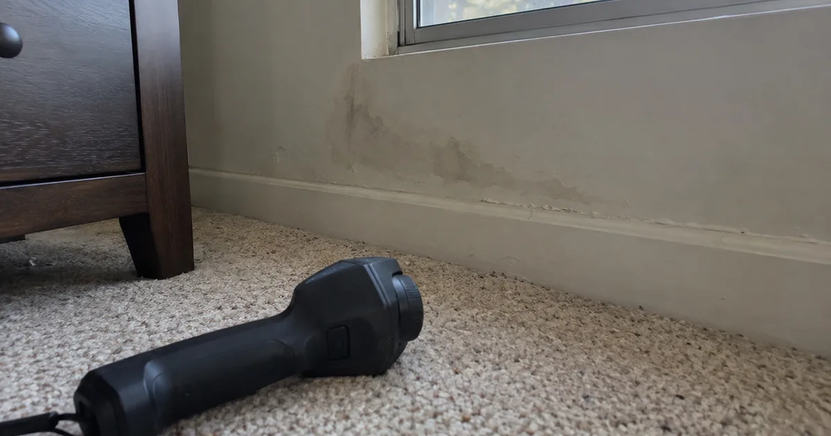 A handheld thermal imaging camera lying on bedroom carpet facing an exterior wall below a window, where a faint watermark and bubbled paint show above the baseboard