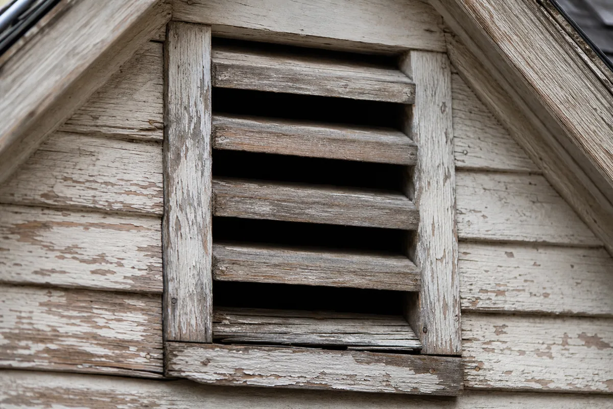 Macro view of an old wooden gable attic vent with wide slats and no fine mesh screening on weathered wood siding