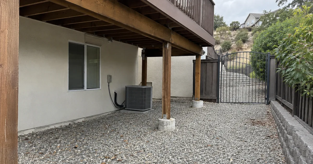 A cleared defensible-space zone under a raised wood deck, gravel covering the ground, a metal gate visible nearby