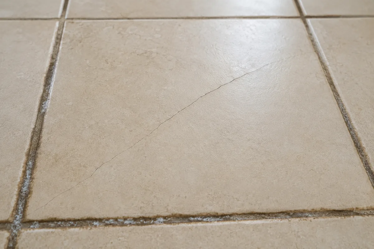 Close-up of a beige porcelain floor tile in a Lake Elsinore home showing a hairline crack running diagonally across the tile, with darkened grout and white mineral efflorescence along the surrounding grout lines, consistent with prolonged moisture from a slab leak underneath