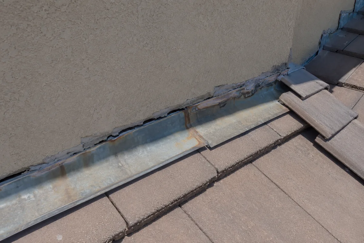 Roof-to-wall flashing failure where stucco meets concrete tile roof showing dried cracked sealant and rust staining on galvanized metal