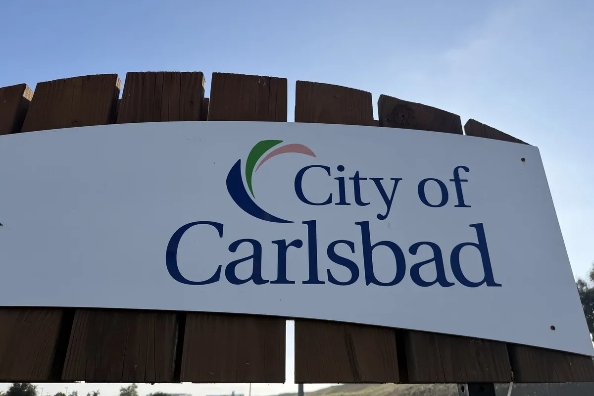 White City of Carlsbad entry sign with blue lettering and a curved logo, mounted on a wood slat frame against blue sky