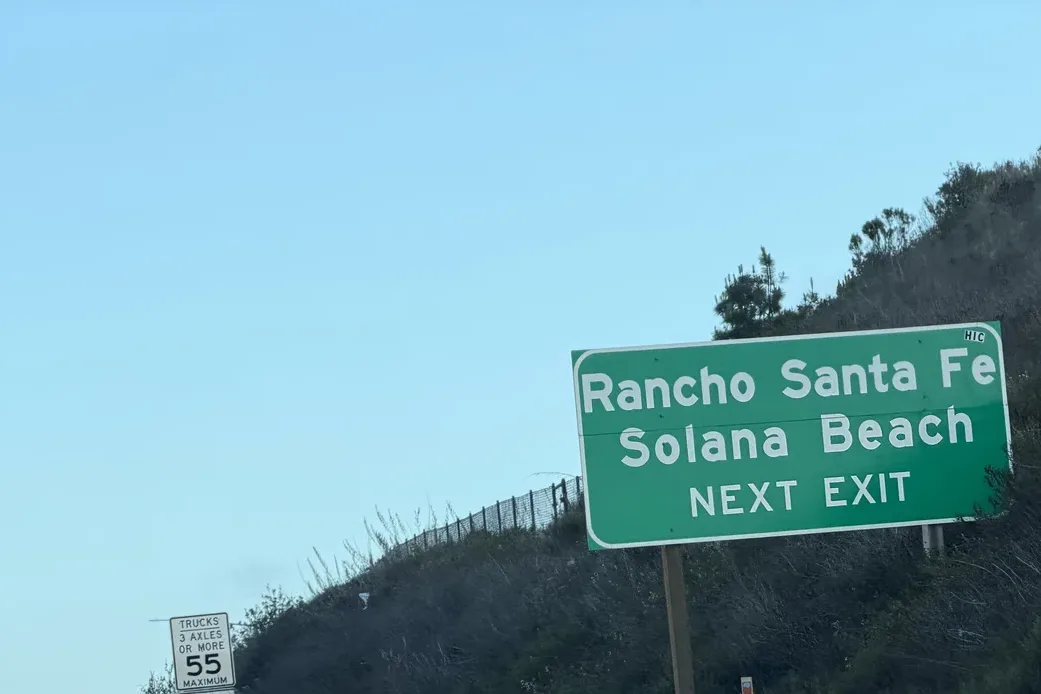 Green freeway sign reading Rancho Santa Fe, Solana Beach, next exit, on a brushy hillside