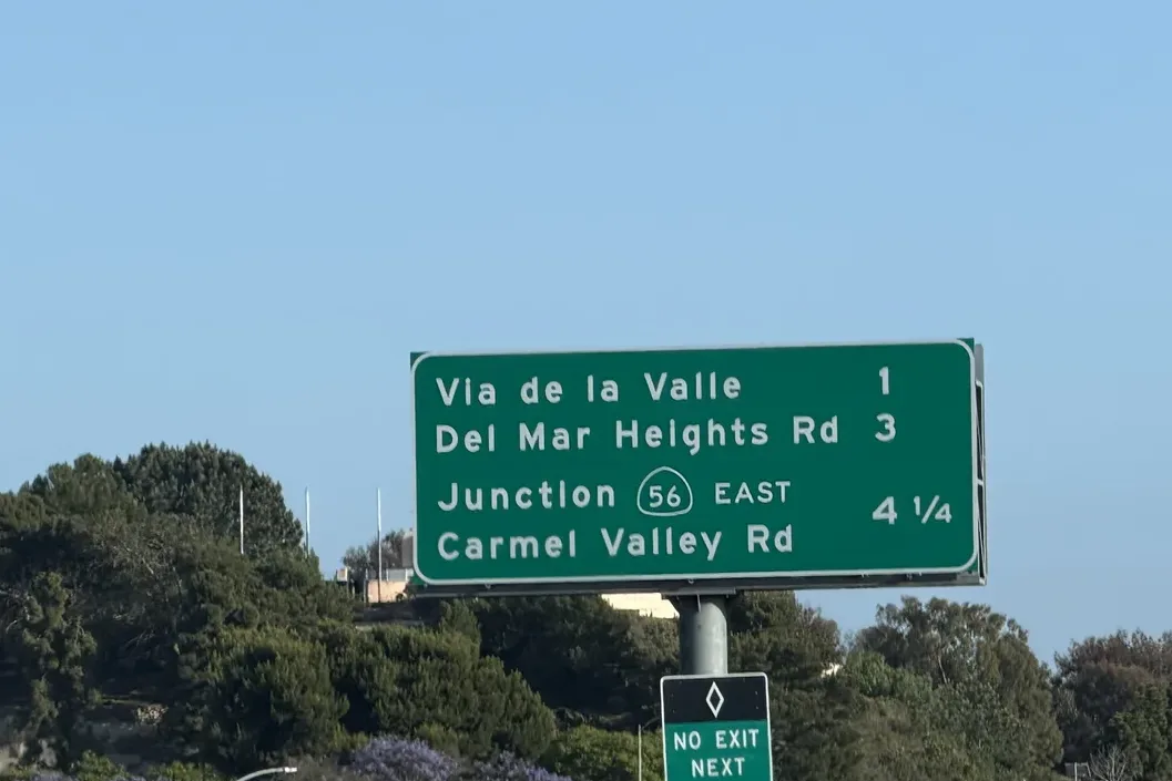 Green freeway sign listing Via de la Valle 1, Del Mar Heights Rd 3 and Junction 56 East Carmel Valley Rd 4 1/4, above coastal trees