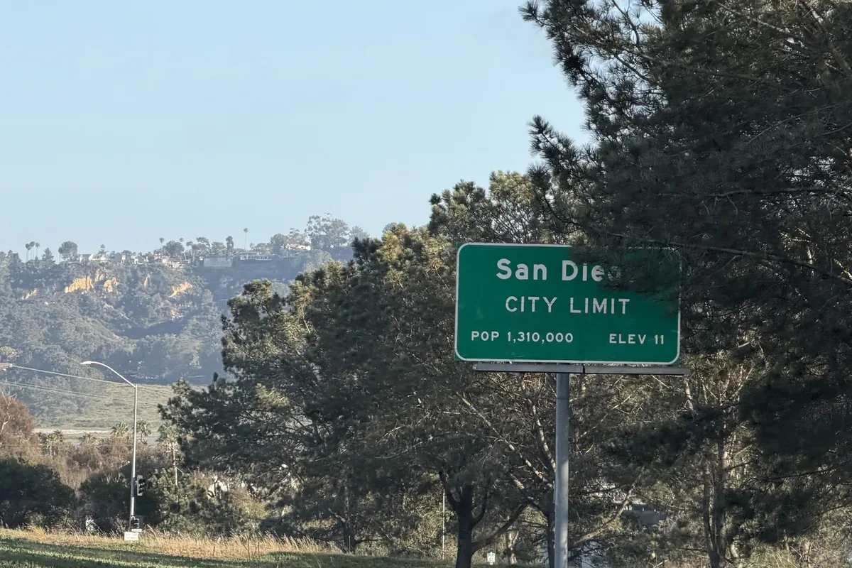 Green San Diego City Limit road sign showing population 1,310,000 and elevation 11, with pine trees and coastal hills behind it