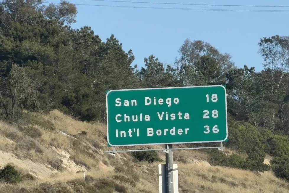 Green roadside mileage sign reading San Diego 18, Chula Vista 28, Int’l Border 36 on a dry hillside with trees