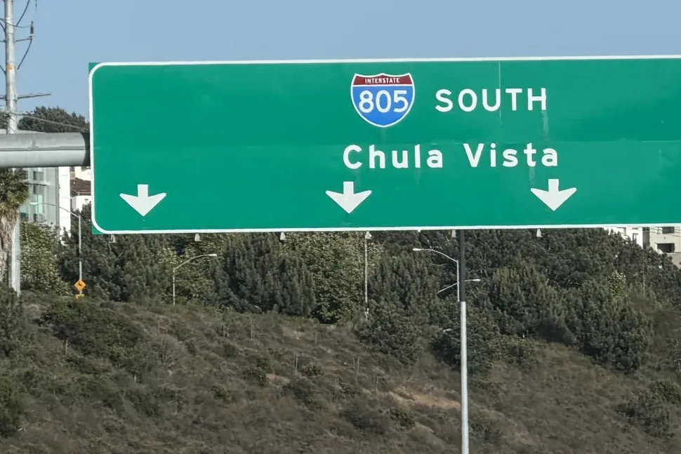 Green overhead freeway sign for Interstate 805 South to Chula Vista with three down arrows above a brushy hillside
