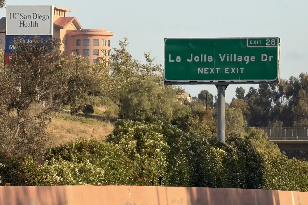 Green La Jolla Village Dr Exit 28 next exit sign above roadside shrubs, with a UC San Diego Health building in the background