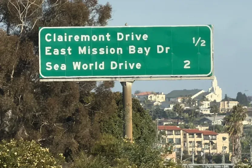 Green overhead freeway sign listing Clairemont Drive, East Mission Bay Dr and Sea World Drive, with hillside homes and a white steeple in the background